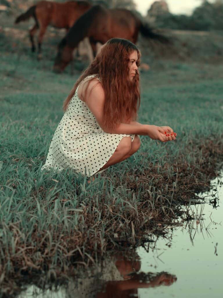 Woman in sleeveless white dress on her haunches on the grass shore of a pond or small lake. The woman's reflection is in the still water with two brown horses in the background.