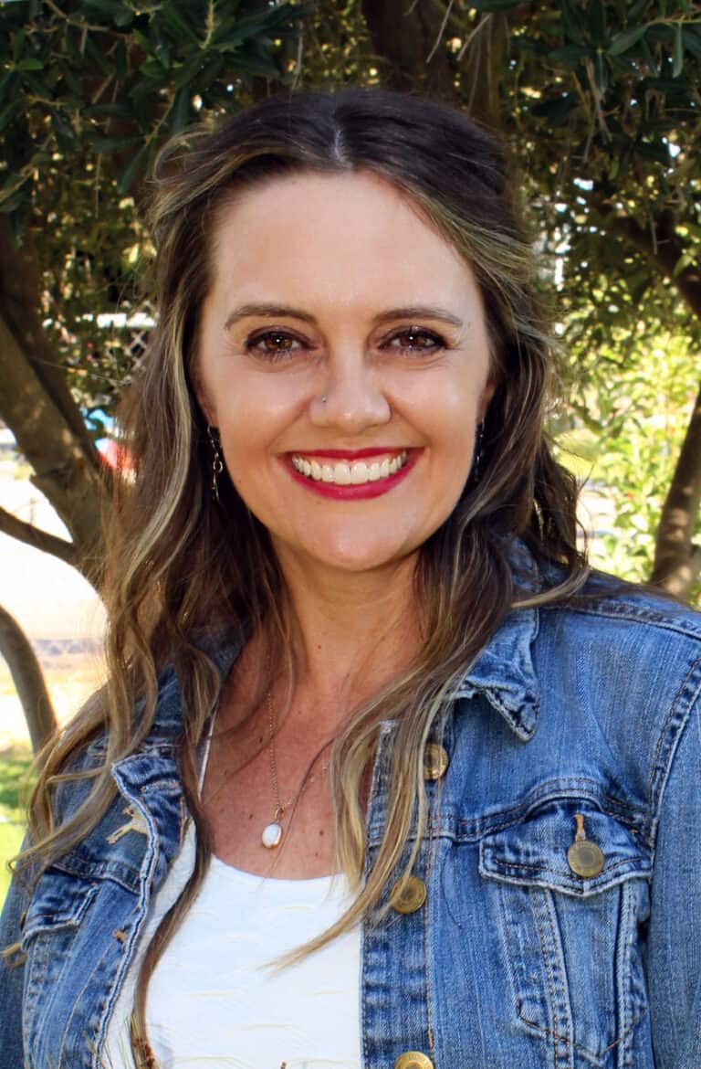 Image of woman in white shirt and jean jacket with mid-length brown hair smiling as she stands outside posing.