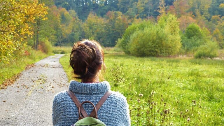 The back of a woman in a light blue sweater and backpack purse, dark hair up in a messy bun, overlooking a pathway along a green meadow with bushes that are beginning to turn fall colors at their tips.